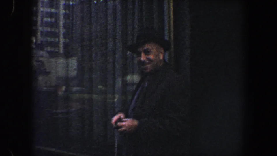 Smiling man in a dark coat and hat faces the camera beside a shadowed New York City building facade.