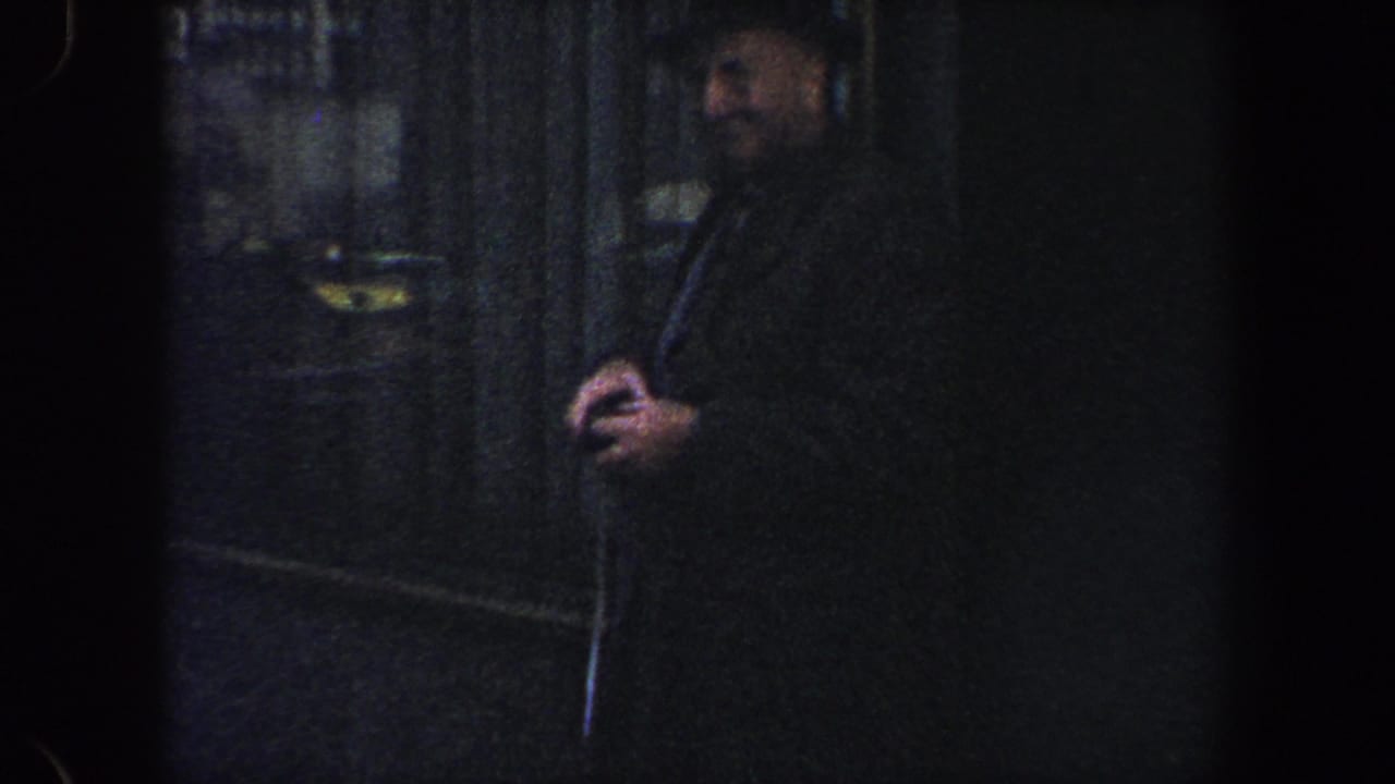 Smiling man in a dark coat and hat standing near a shadowed city doorway.