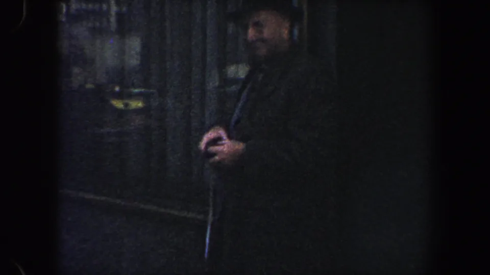 A smiling man in a dark coat and hat stands close to the camera on a shadowed New York City street in 1960s home-movie footage.