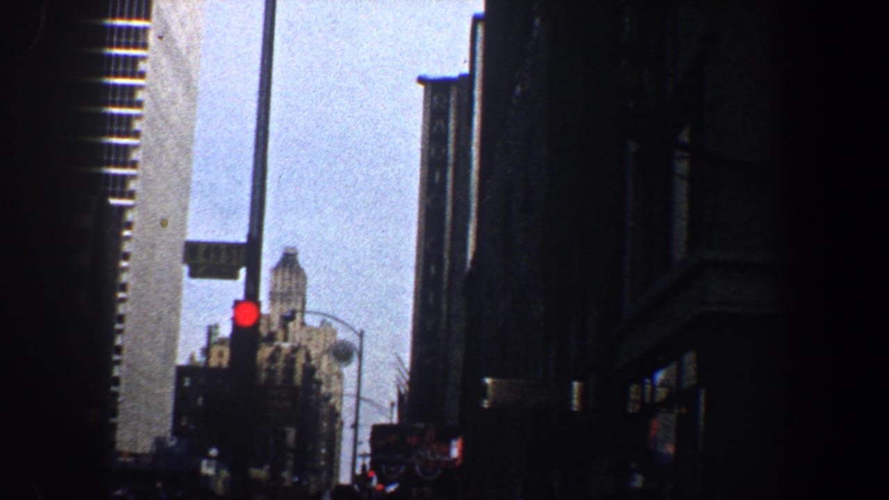 Tall city buildings frame a street view with a red traffic light and pale sky.