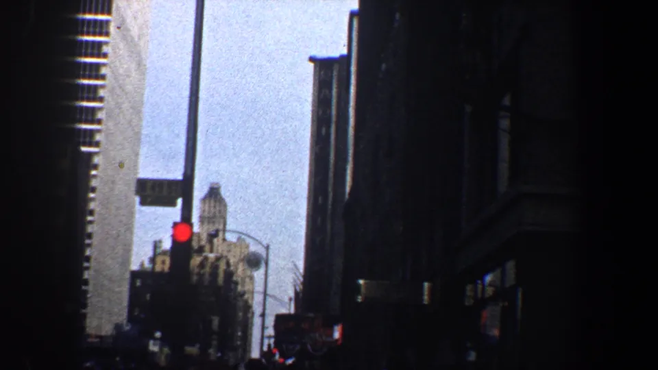 Tall New York City buildings frame a street view with a red traffic light, distant architecture, and deep shadows from 1960s 8mm footage.