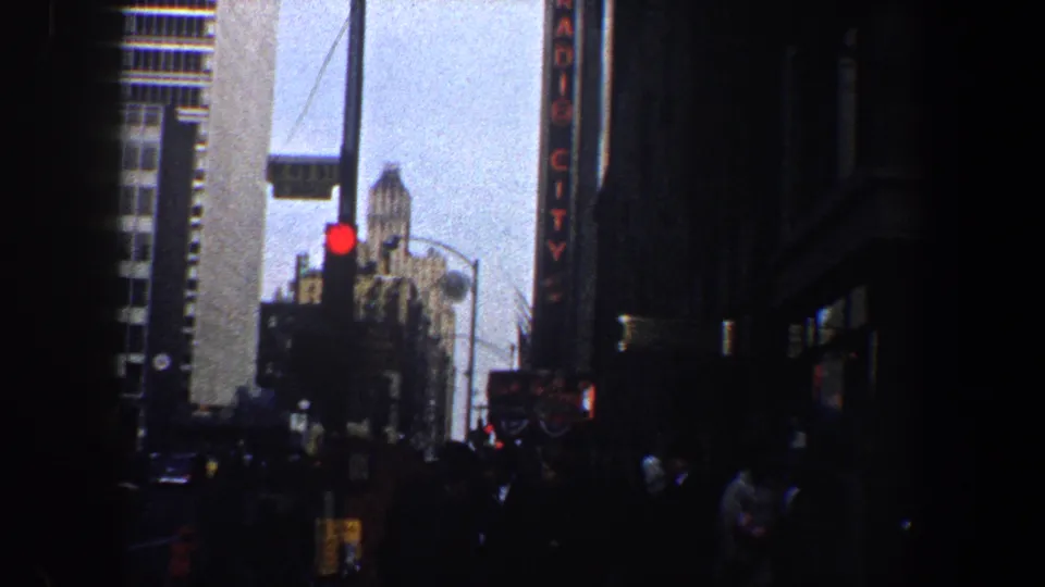 A busy New York City street view shows pedestrians, cars, a red traffic light, and part of the vertical RADIO CITY sign in 1960s footage.