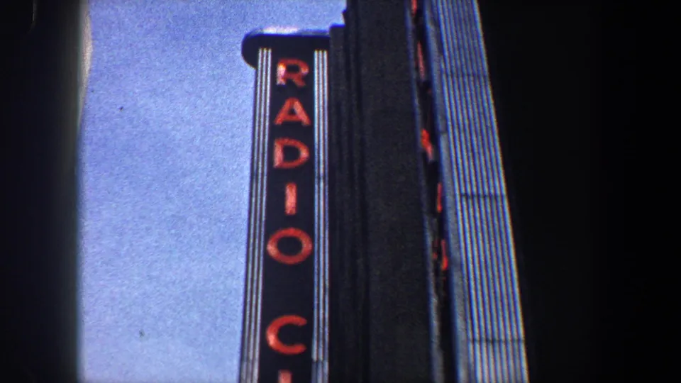 A vertical RADIO CITY sign fills the frame beside a tall building, with blue sky and shadowed architecture in 1960s New York footage.