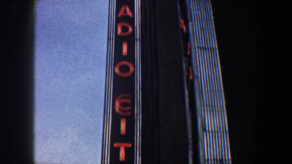 1960 New York City view of the Radio City vertical neon sign rising beside a dark Art Deco facade.
