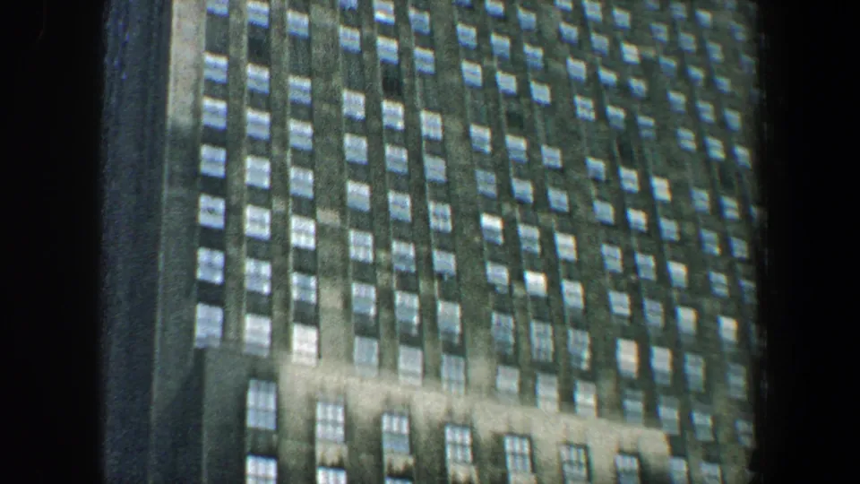 Upward-looking 8mm frame of a New York City high-rise at night, with repeated window rows and dark framing at the side.