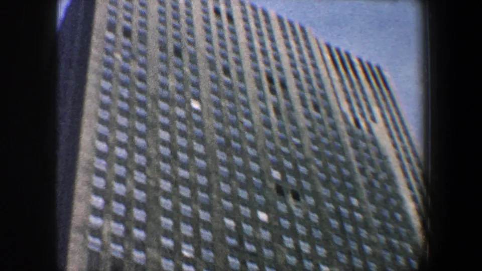 1960 New York City 8mm frame looking up at the upper stories of a gridded business building against a pale blue sky.