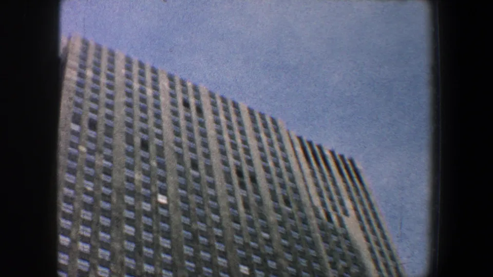 1960 New York City 8mm frame of a high-rise edge and repeated windows, tilted upward from street level into blue sky.