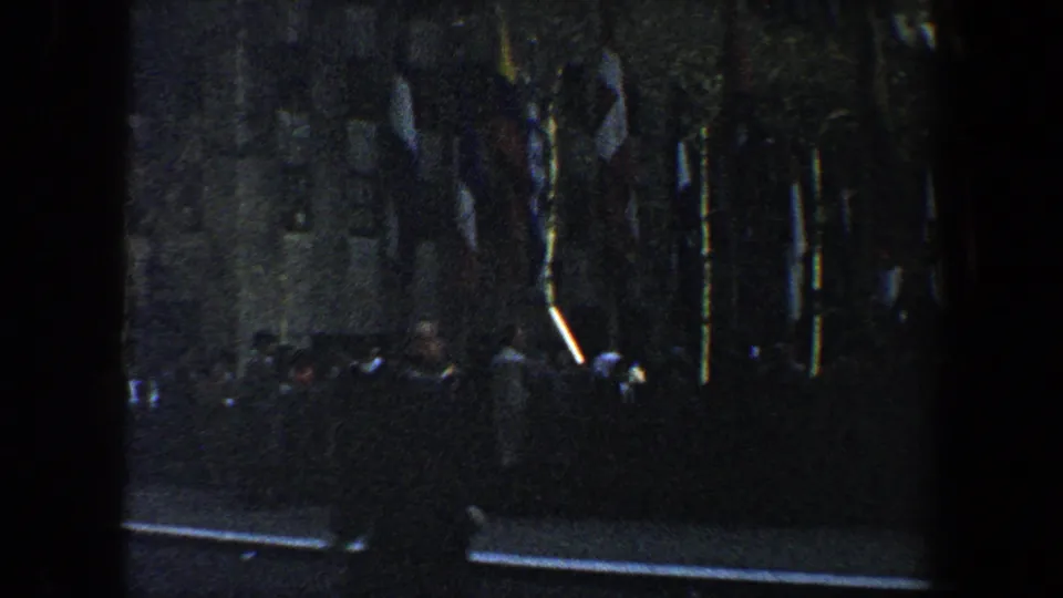 1960 New York City 8mm frame dropping to a shadowed street-level area with several flags near the base of a building.