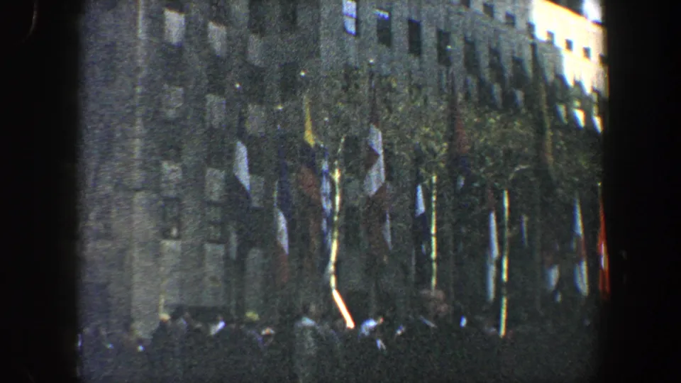 1960 New York City 8mm frame showing people at street level beneath a large flag-lined building facade.