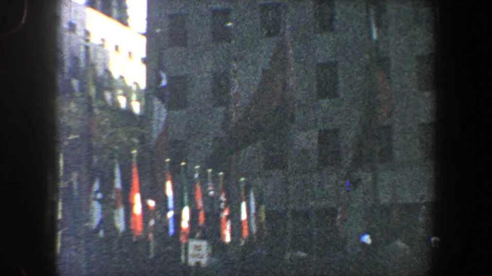 Low-light 1960 New York City frame with flags along the street edge and people gathered near a large building.
