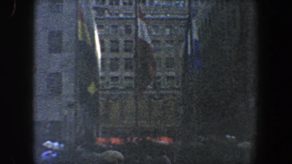 1960 New York City 8mm frame showing a shadowed public crowd, indistinct flags, and tall urban building facades at night.