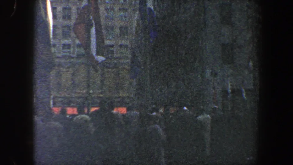 1960 New York City nighttime frame with people gathered below indistinct flags, trees, and multi-story urban buildings.