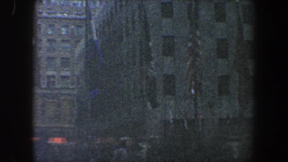 Low-light 8mm view of a New York City crowd edge, tall facade details, and faint flags during a 1960 public gathering.