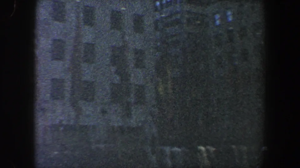 Dark 1960 New York City home-movie frame showing a public crowd, trees, and building facades partly obscured by 8mm frame edges.