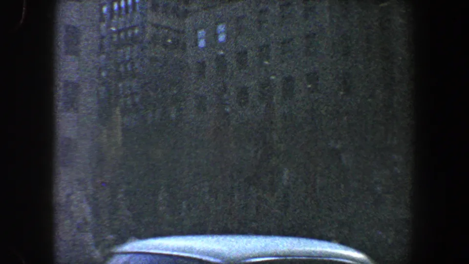 1960 New York City 8mm frame showing a dark downtown building facade, scattered lit windows, rain texture, and the roof of a car at the bottom edge.