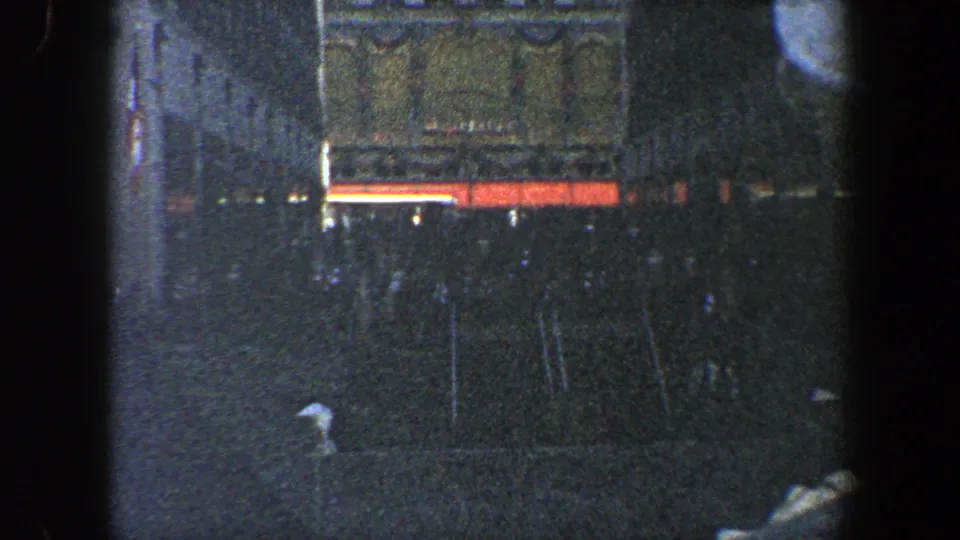 1960 New York City 8mm frame with a dark high-rise facade, bright horizontal street-level lights, rain streaks, and indistinct movement near the sidewalk.