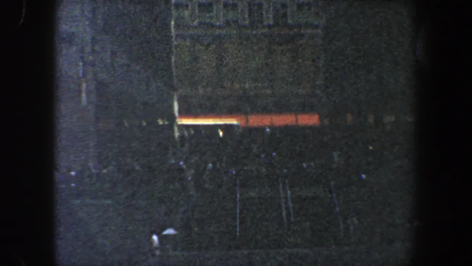 1960 New York City 8mm frame showing a rain-darkened building, glowing street-level lights, indistinct sidewalk forms, and a partial car edge at the bottom.