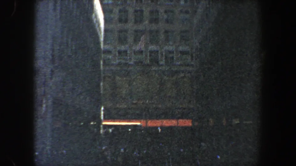 1960 New York City 8mm frame with a shadowed building wall, rows of windows, a bright vertical strip, and red-orange lights near street level.