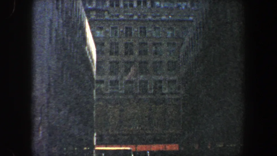 Low-angle 1960 New York City street view framed by dark building edges and a hazy high-rise facade on 8mm film.