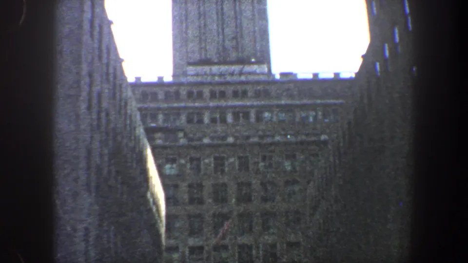 Upward-looking 1960 New York City view of a tower roofline and adjacent building walls against a bright hazy sky.