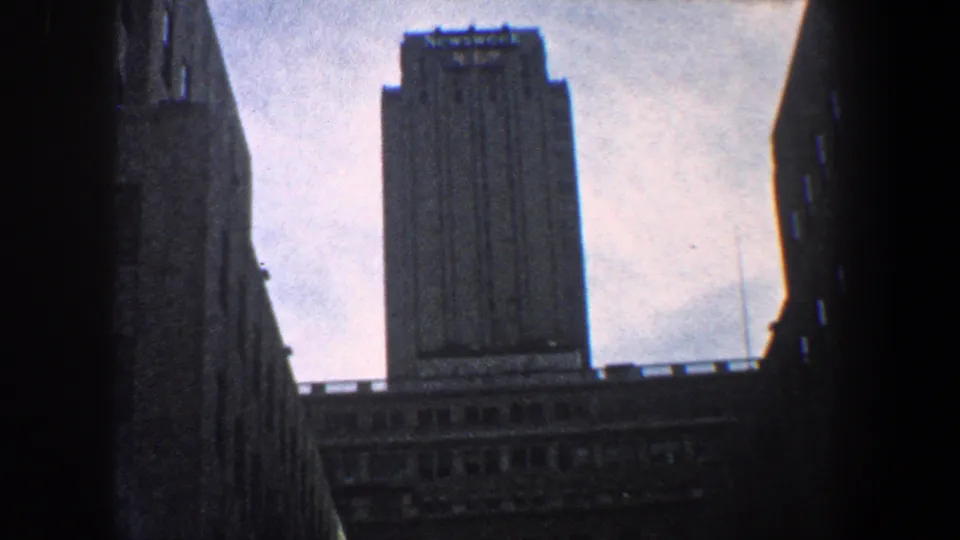 Street-level 1960 New York City view looking up toward a tall tower, with surrounding buildings forming a narrow vertical frame.