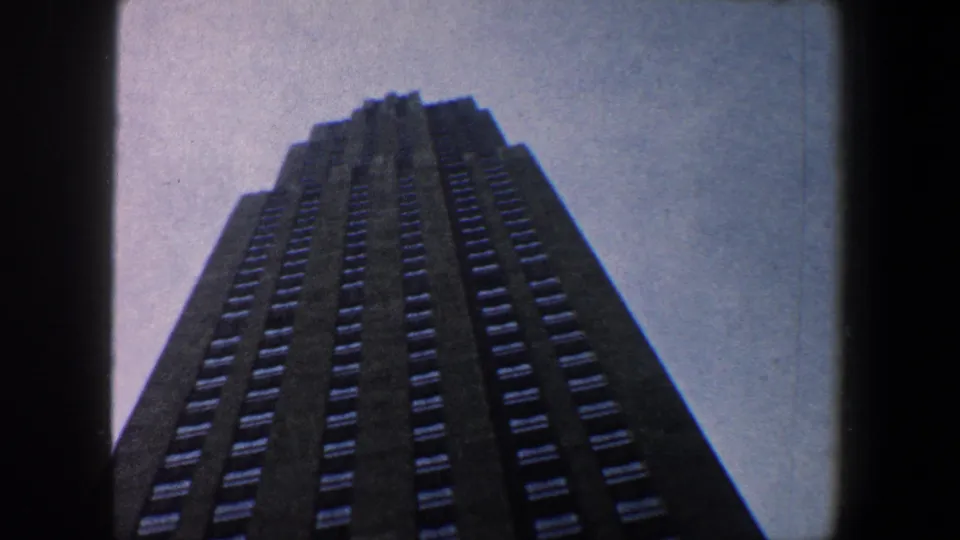 A steep low-angle 1960 New York City shot of a dark skyscraper facade with repeating window rows against a pale sky.