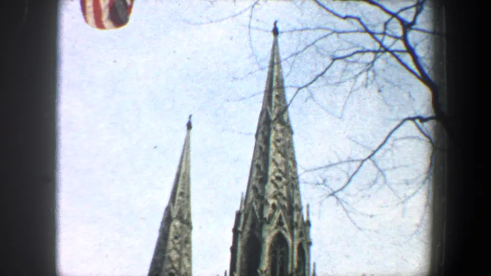 Bright 1960 New York City frame with Gothic church spires, bare tree branches, pale sky, and a partial American flag.