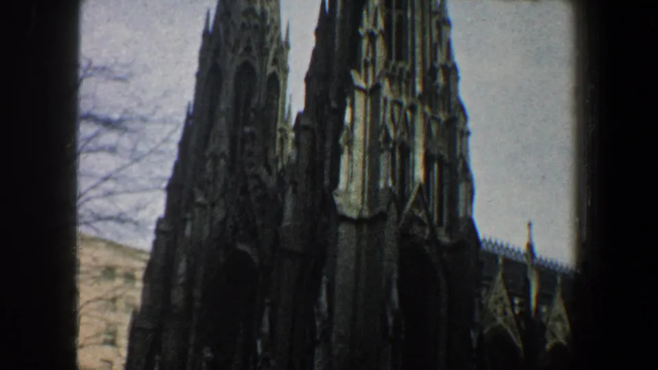 1960 New York City 8mm frame showing the church spires and side facade, with bare branches and a neighboring building edge visible.