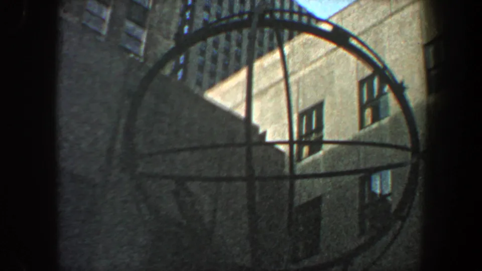 Low-angle 1960 New York City 8mm view of a circular metal sculpture with tall building facades behind it.