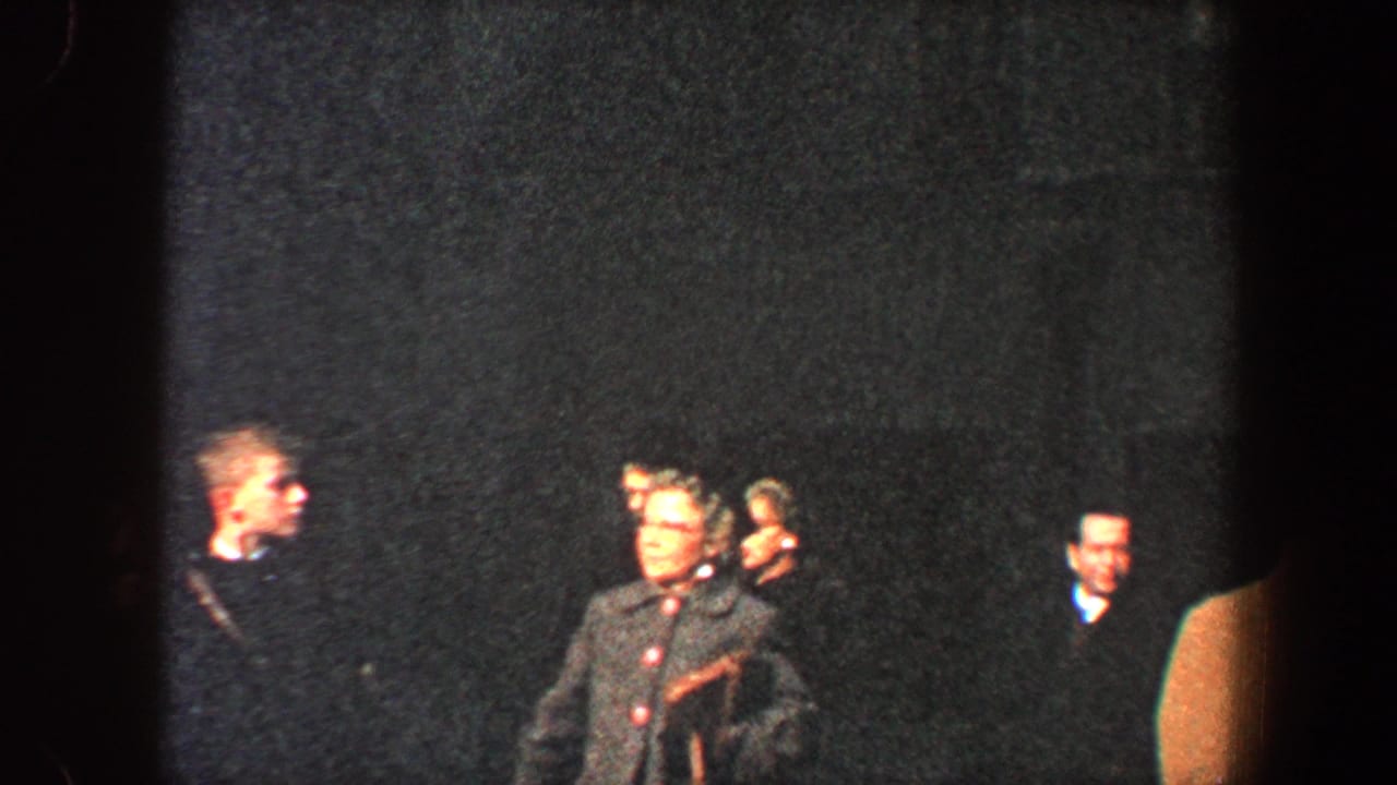 Several adults stand or walk in low light, including a woman in a dark coat and a suited man.