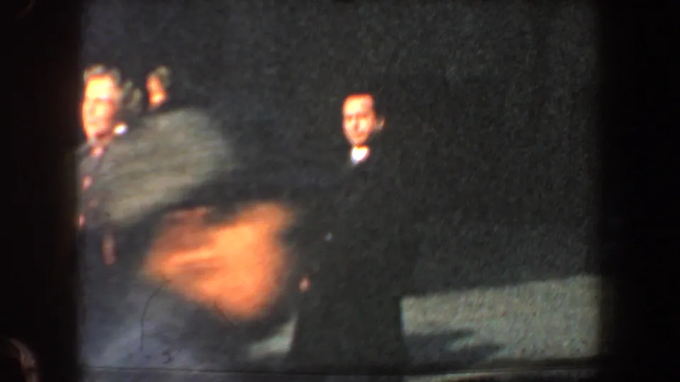 Low-light 1960 New York City frame with a blurred foreground passerby and background adults near a dark wall.