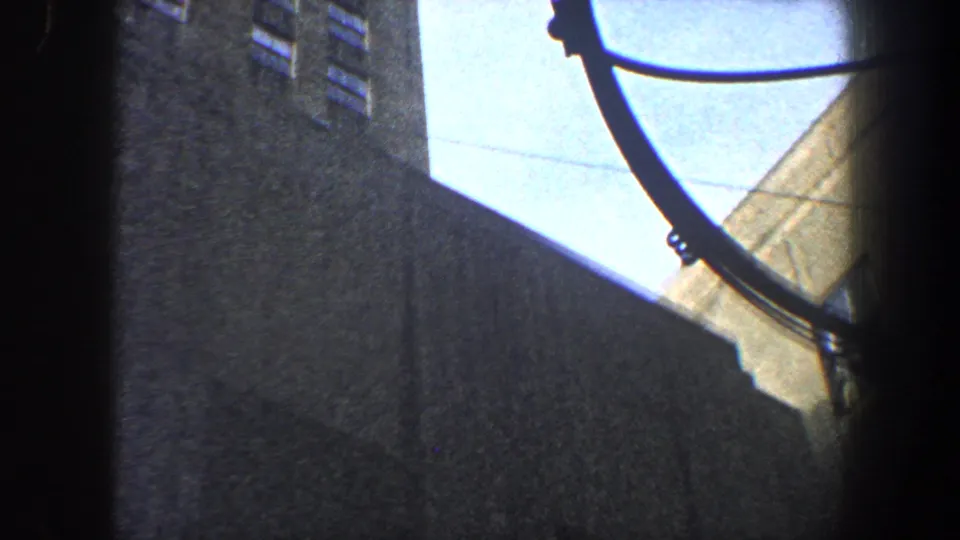 Grainy 1960 New York City home-movie frame focused on a bright building edge, deep shadow, and curved dark line.