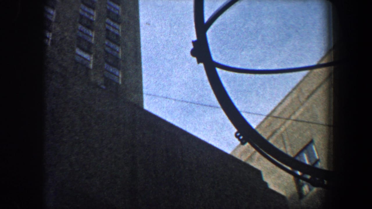 1960 New York City 8mm frame looking upward between urban buildings toward part of an ornate high-rise facade and a circular street fixture.