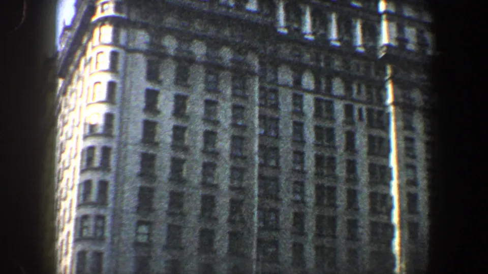 1960 New York City 8mm frame showing the building corner from below, with a rounded bay, tall window rows, and sunlit stonework.