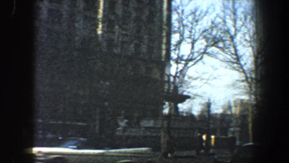 1960 New York City street view from a moving vehicle with bare trees, traffic, and a tan building partly obscured by dark framing.