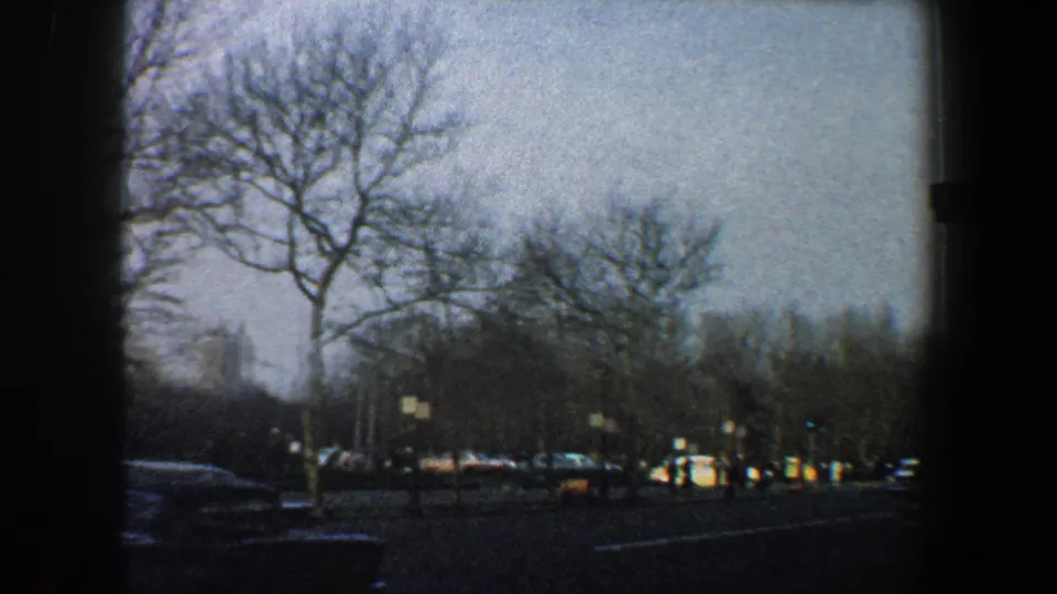 1960 New York City road view with bare trees, light traffic, and low city buildings captured in grainy 8mm color.