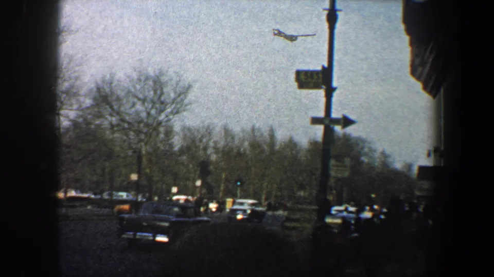 New York City street corner view with cars, bare trees, a street sign, traffic signals, and a small aircraft overhead.