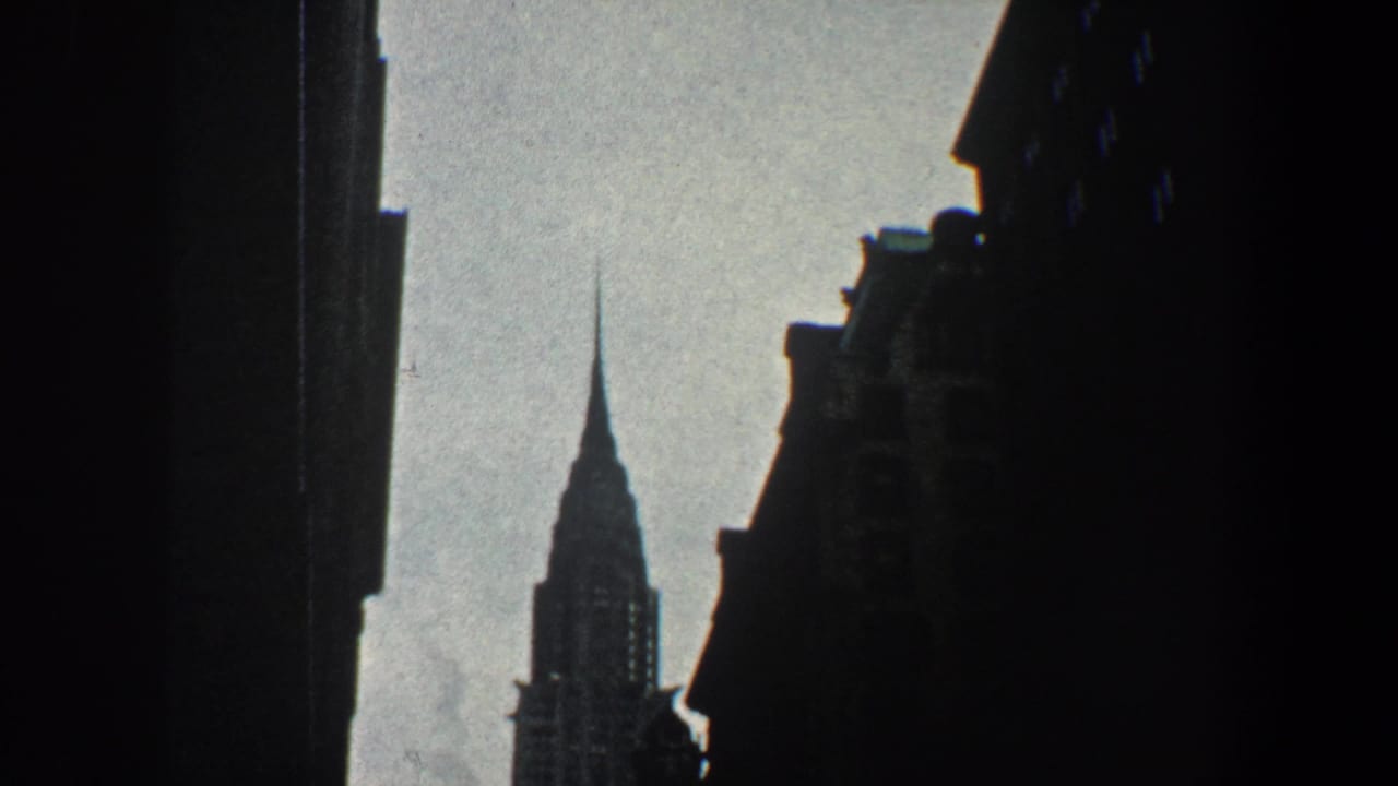 Chrysler Building spire seen between dark building silhouettes against a pale sky.