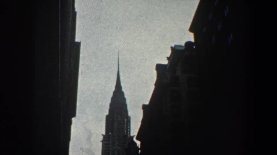 The Chrysler Building rises in the distance between dark Manhattan building edges in 1960 8mm footage.