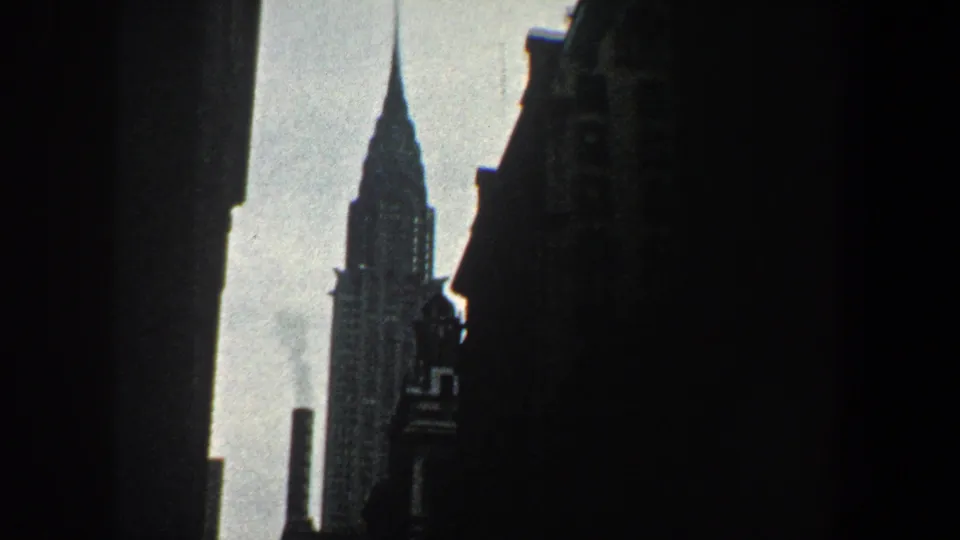 1960 New York City 8mm frame showing the Chrysler Building spire framed by dark building silhouettes and bright sky.