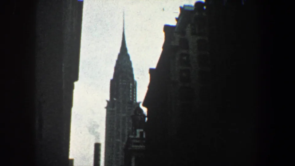 1960 New York City frame centering the Chrysler Building spire between silhouetted buildings with faint smoke against the pale sky.