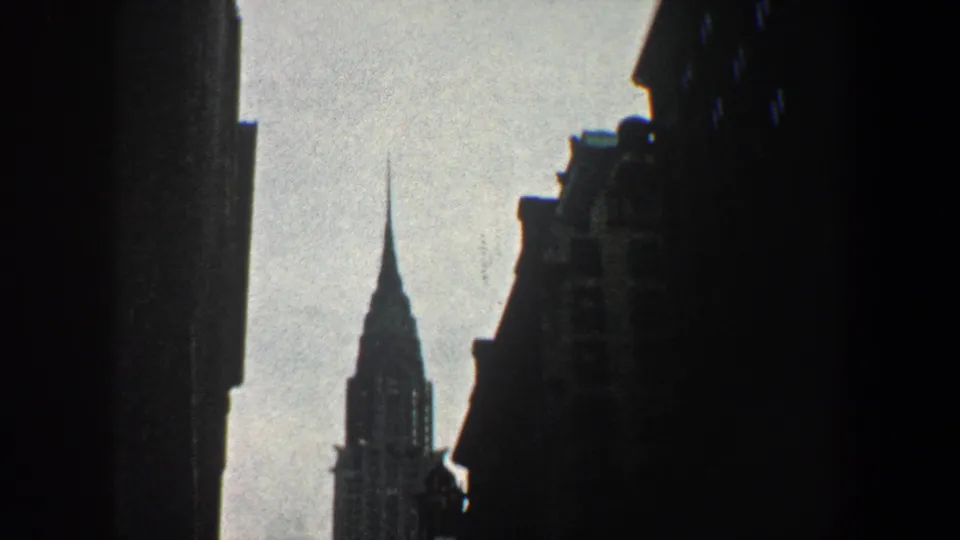 1960 New York City view of the Chrysler Building spire rising between shadowed urban buildings.