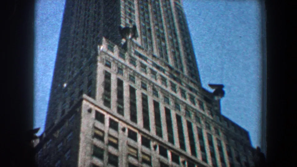 1960 New York City 8mm frame showing the upper portion of a tall Art Deco-style skyscraper tapering upward against blue sky.