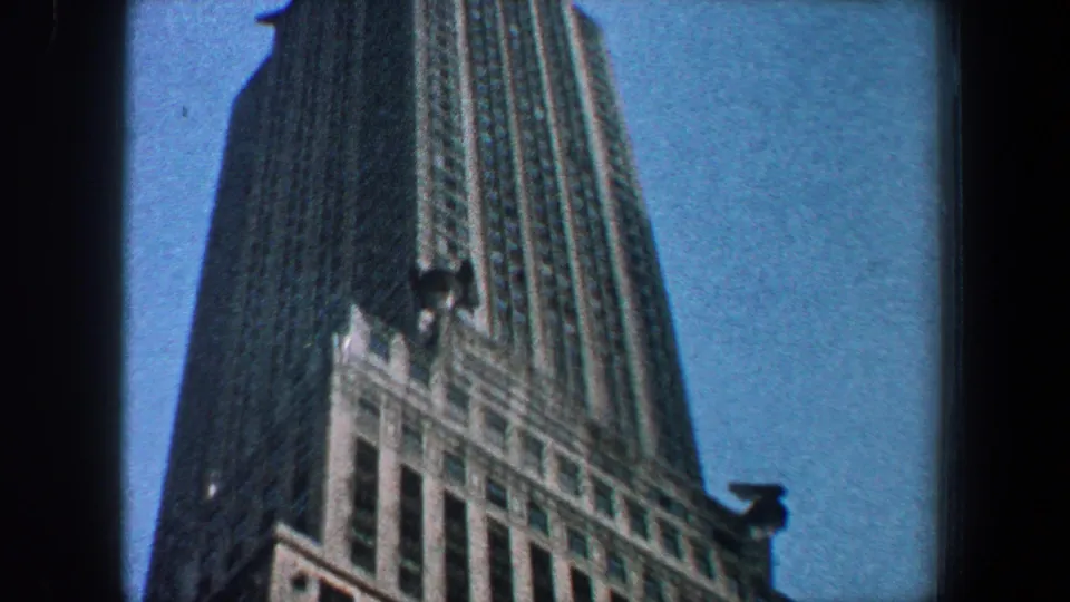 1960 New York City home-movie frame showing the Empire State Building from below, with its facade and upper tower against blue sky.