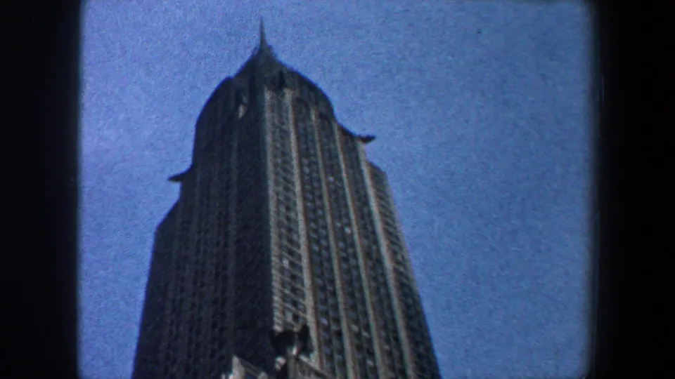 1960 New York City frame focusing on the Empire State Building's upper tower and spire, bordered by dark 8mm film edges.