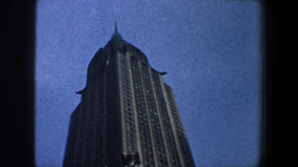 1960 New York City home-movie frame with the Empire State Building tower seen from below, slightly darker and framed by film edges.