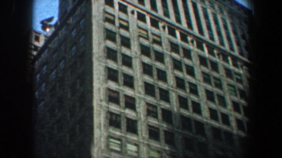 1960 New York City home-movie frame showing a low-angle view of a tall office building facade with rows of windows.