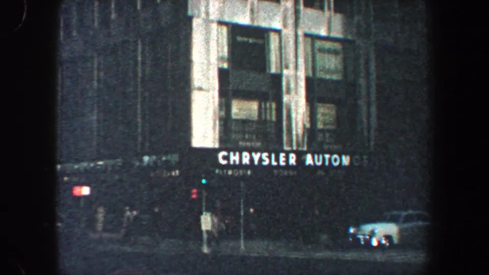 1960 New York City street-level frame showing the Chrysler Automotive sign beneath the facade, with traffic lights and a car nearby.