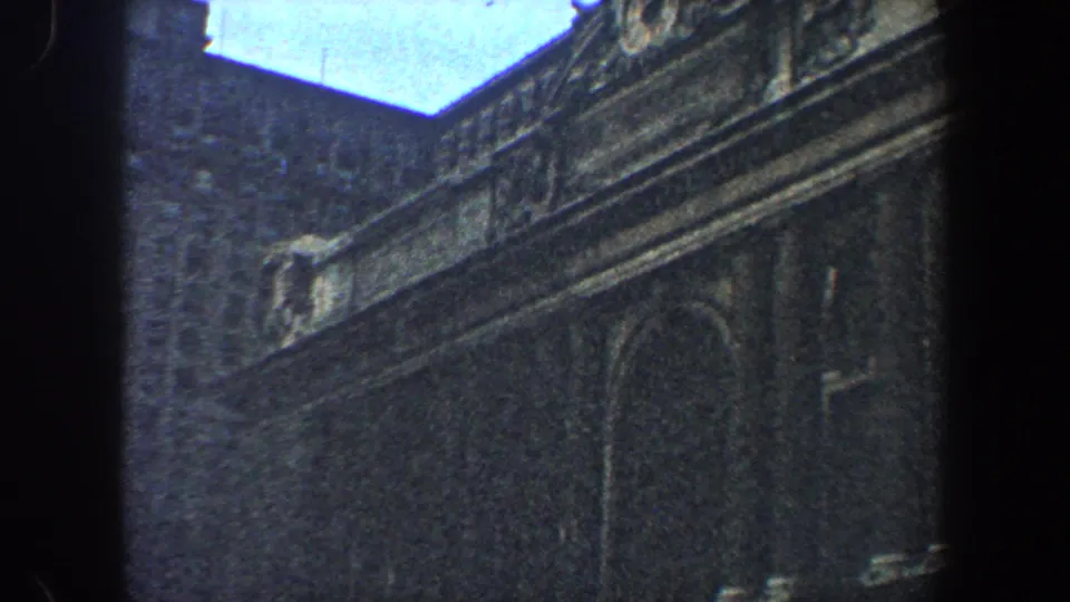 Visitor-shot 1960 New York City frame looking up at a historic stone facade with carved trim, arched window outlines, and sky at the roofline.
