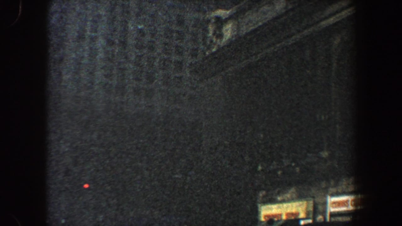 1960 New York City 8mm frame looking up at shadowed skyscraper walls, with small storefront signs near the lower edge.